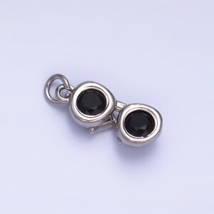 May include: A silver-colored charm in the shape of sunglasses, featuring two black, round gemstones for lenses. The charm has a small loop at the top for attaching to a necklace or bracelet. The design is simple and elegant.