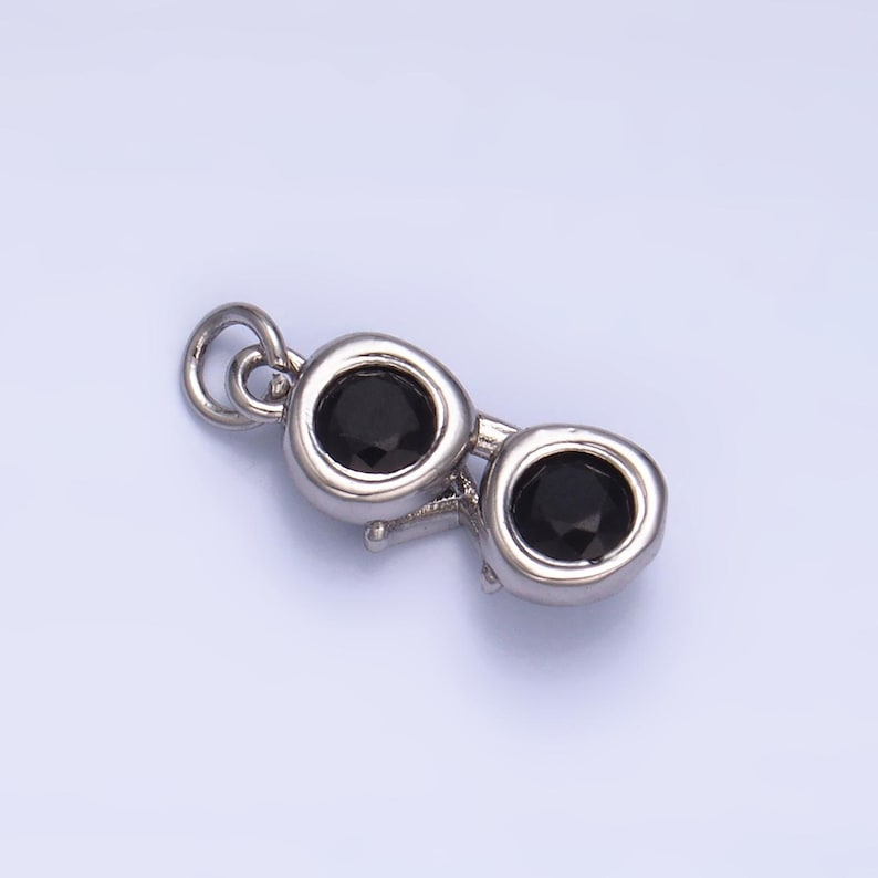 May include: A silver-colored charm in the shape of sunglasses, featuring two black, round gemstones for lenses. The charm has a small loop at the top for attaching to a necklace or bracelet. The design is simple and elegant.