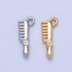 Puede incluir: Dos pequeños colgantes de peine, uno plateado y otro dorado, se muestran sobre un fondo morado claro. Cada peine tiene un mango con un diseño floral y un lazo en la parte superior para sujetar.