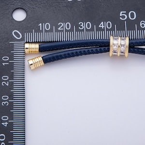 May include: A navy blue leather cord necklace with gold-colored metal accents. The necklace features a central gold-colored bead with clear rectangular stones. The necklace is shown against a white background.
