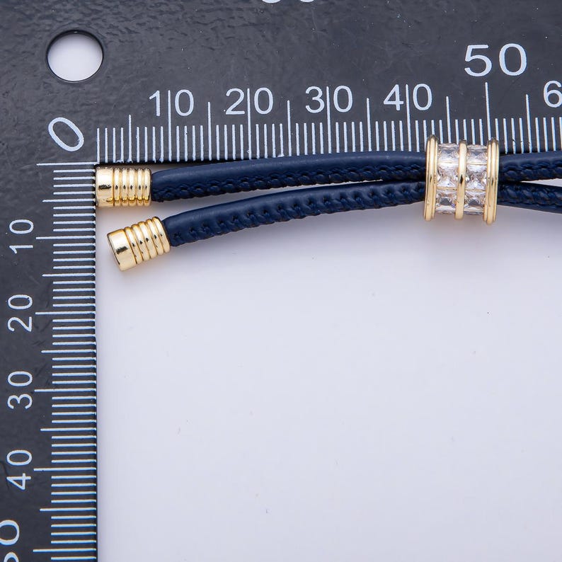 May include: A navy blue leather cord necklace with gold-colored metal accents. The necklace features a central gold-colored bead with clear rectangular stones. The necklace is shown against a white background.