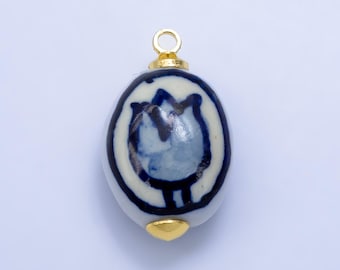 May include: A ceramic pendant with a blue floral design on a white background. The oval-shaped pendant has a gold-colored loop at the top and a gold-colored cap at the bottom. The design is reminiscent of traditional blue and white pottery.