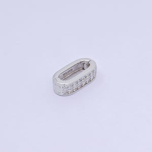 May include: A silver-colored oval clasp with a row of small, clear stones set along the edges. The clasp is designed to open and close, and it is set against a white background.