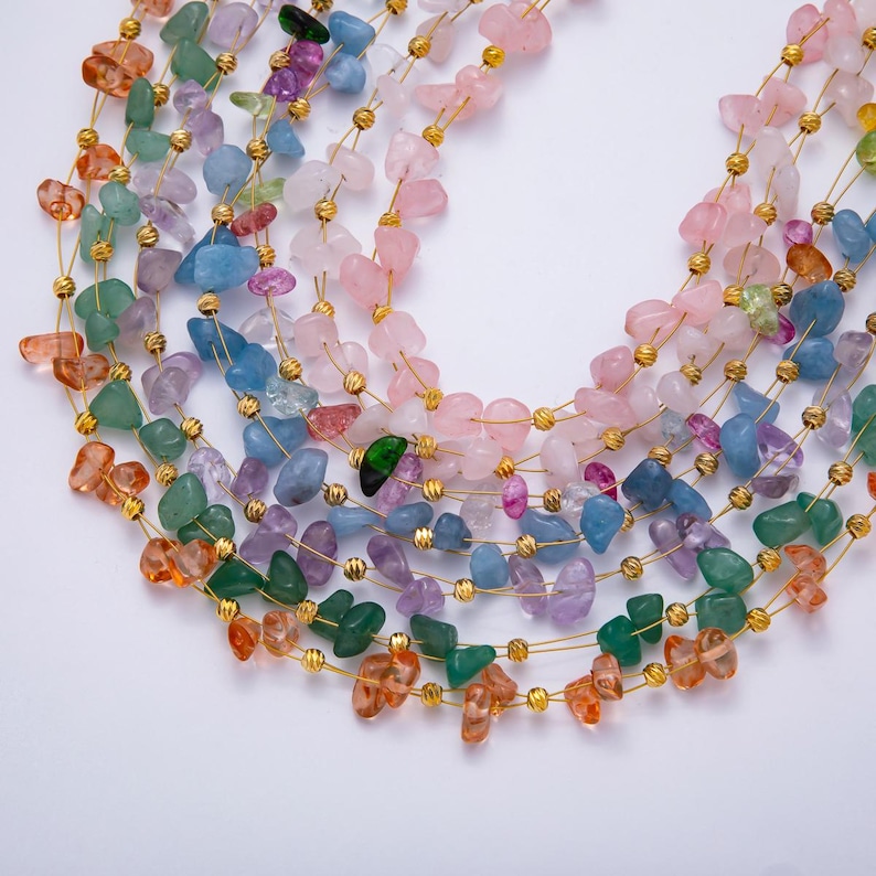 May include: A collection of colorful gemstone necklaces. Each necklace features a strand of irregularly shaped stones in shades of pink, blue, green, orange, and purple, strung on gold-colored wire. The necklaces are arranged in a layered, cascading design.