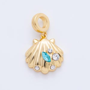 Puede incluir: Un charm dorado con forma de concha, adornado con tres cristales transparentes y un cristal azul claro de forma ovalada. El charm tiene un lazo en la parte superior para sujetarlo a una pulsera o collar.