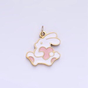 Op de afbeelding: Een gouden hanger in de vorm van een wit konijn dat een roze hart vasthoudt. De hanger heeft een lus aan de bovenkant om aan een ketting of armband te bevestigen.