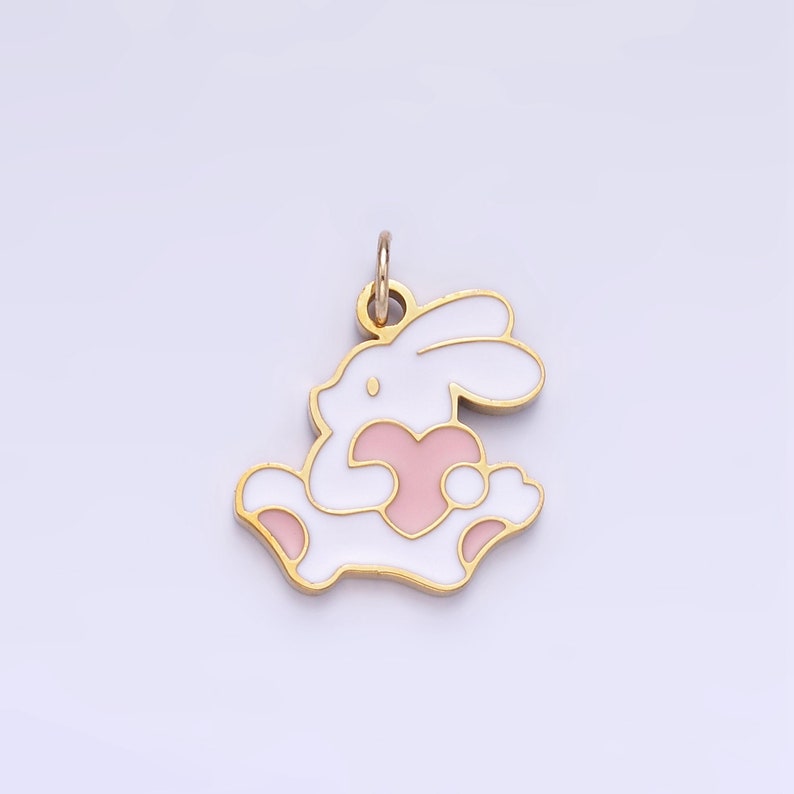 Op de afbeelding: Een gouden hanger in de vorm van een wit konijn dat een roze hart vasthoudt. De hanger heeft een lus aan de bovenkant om aan een ketting of armband te bevestigen.