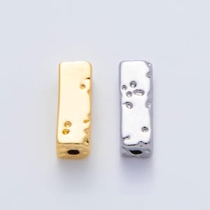 May include: Two rectangular metal beads with textured surfaces. One bead is gold-colored, the other is silver-colored. Both have a hole at the bottom. The beads are likely for jewelry making or crafting.