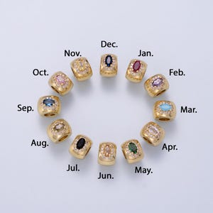 May include: A collection of twelve gold-toned metal beads arranged in a circle, each featuring a central oval-shaped colored gemstone surrounded by small clear crystals. Each bead is labeled with a month abbreviation from Jan. to Dec.