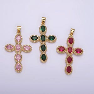 May include: Three gold cross pendants with different colored gemstones. The cross on the left has pink gemstones, the middle cross has green gemstones, and the cross on the right has red gemstones.