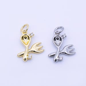 May include: Two pendant charms, one gold-toned and one silver-toned, each featuring a spoon and fork design. Both charms have a small loop at the top and are embellished with clear, sparkling accents. The charms are set against a white background.