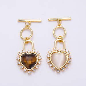 May include: Two gold-toned metal pendants featuring a heart-shaped center stone surrounded by small clear crystals. One heart is dark brown with a striped pattern, and the other is a smooth, opaque off-white. Both hang from a gold toggle bar.