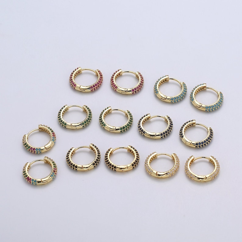 Pu&ograve; includere: Una collezione di orecchini a cerchio dorati, ciascuno ornato da una fila di pietre preziose colorate. Gli orecchini presentano vari colori di pietre preziose, tra cui rosso, verde, blu e nero. Gli orecchini sono esposti su una superficie bianca.