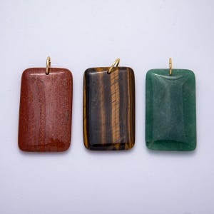 May include: Three rectangular gemstone pendants with gold-colored loops. The pendants are red, brown with yellow stripes, and green. The gemstones are polished and have a smooth surface. The pendants are likely for necklaces or other jewelry.