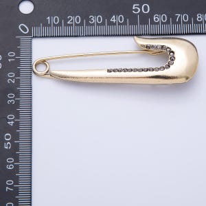 May include: A gold-colored safety pin with a row of small, dark-colored stones along the pin's body and head. The pin is shown against a white background with a ruler in the upper left corner.