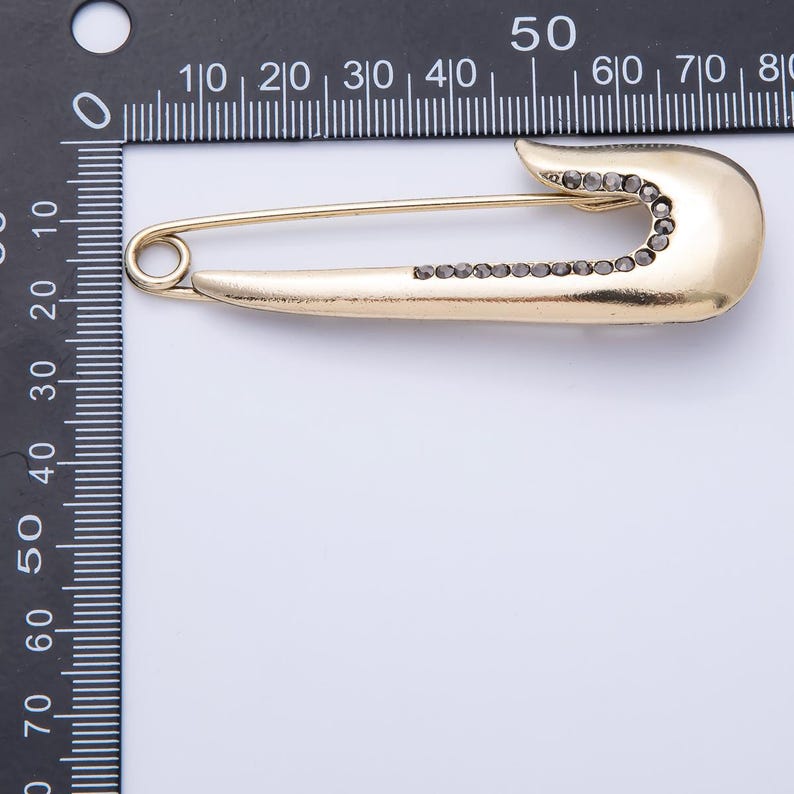 May include: A gold-colored safety pin with a row of small, dark-colored stones along the pin's body and head. The pin is shown against a white background with a ruler in the upper left corner.