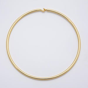 May include: A circular gold-toned metal choker necklace with a smooth, polished finish. The necklace features a simple hook-and-eye style closure mechanism at the top center, creating a continuous, minimalist hoop design for the neck.