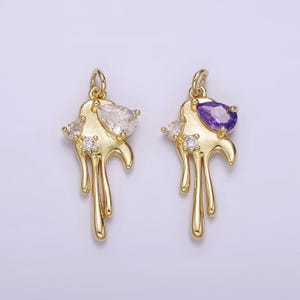 May include: Two gold-colored pendants with a dripping design. One pendant features a clear, pear-shaped gem and small clear accent stones. The other pendant has a purple, pear-shaped gem and clear accent stones. Both have a loop at the top.