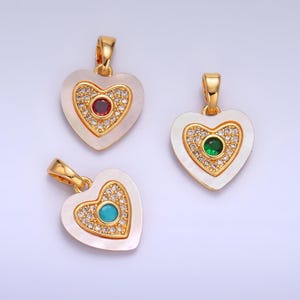 Op de afbeelding: Drie gouden hartvormige hangers met witte parelmoer inlegwerk. Elke hanger heeft een edelsteen in een andere kleur, omringd door heldere strass steentjes.