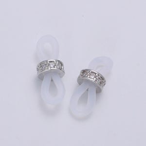 Puede incluir: Dos soportes para gafas de silicona blanca con una banda plateada de diamantes de imitaci&oacute;n.