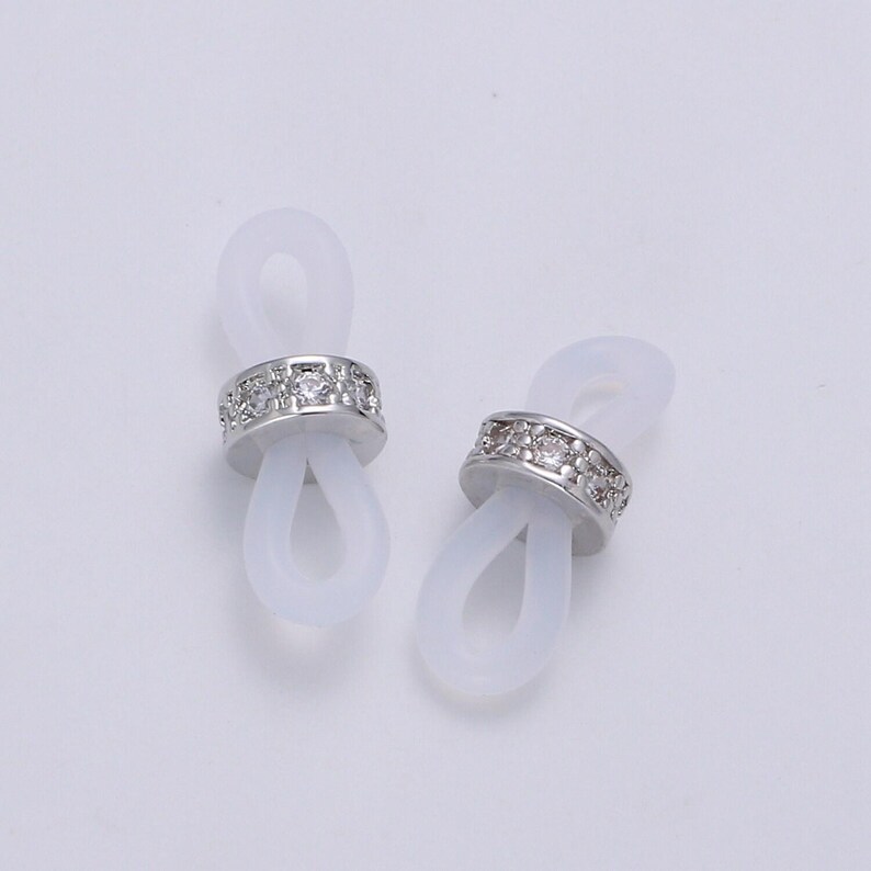 Puede incluir: Dos soportes para gafas de silicona blanca con una banda plateada de diamantes de imitaci&oacute;n.