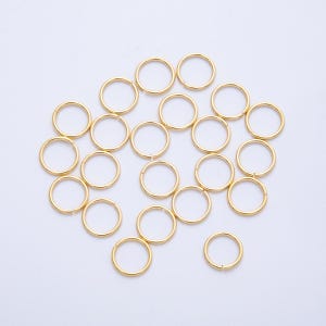 May include: A collection of small, gold-colored jump rings scattered across a white surface. The rings are circular and appear to be made of metal, likely for jewelry making or crafting. The lighting highlights their smooth, reflective surfaces.