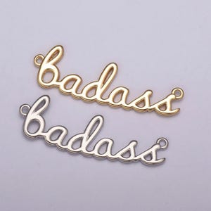 Op de afbeelding: Twee metalen bedels met het woord "badass" geschreven in een cursief schrift. De bovenste bedel is goud en de onderste is zilver.