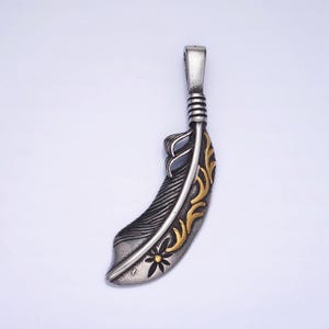 Peut inclure: Un pendentif en forme de plume argentée et dorée avec un design détaillé. La plume est stylisée avec un motif floral et une petite fleur en bas.