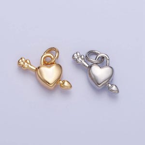 May include: Two heart-shaped charms, one gold-colored and one silver-colored, each pierced by an arrow. The charms have a loop at the top for hanging. The arrow has a decorative tip and fletching. The background is a light purple color.