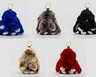May include: Five plush bunny keychains in various colors: blue, gray, black, tan, and red. Each bunny has a gold-colored ring and a leather strap. The bunnies have white paws and a fluffy texture.