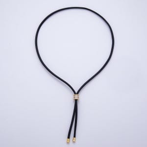 May include: A black cord necklace with a gold-colored central bead encrusted with small, clear stones. The necklace has two dangling black cords with gold-colored tips. The necklace is displayed against a white background.