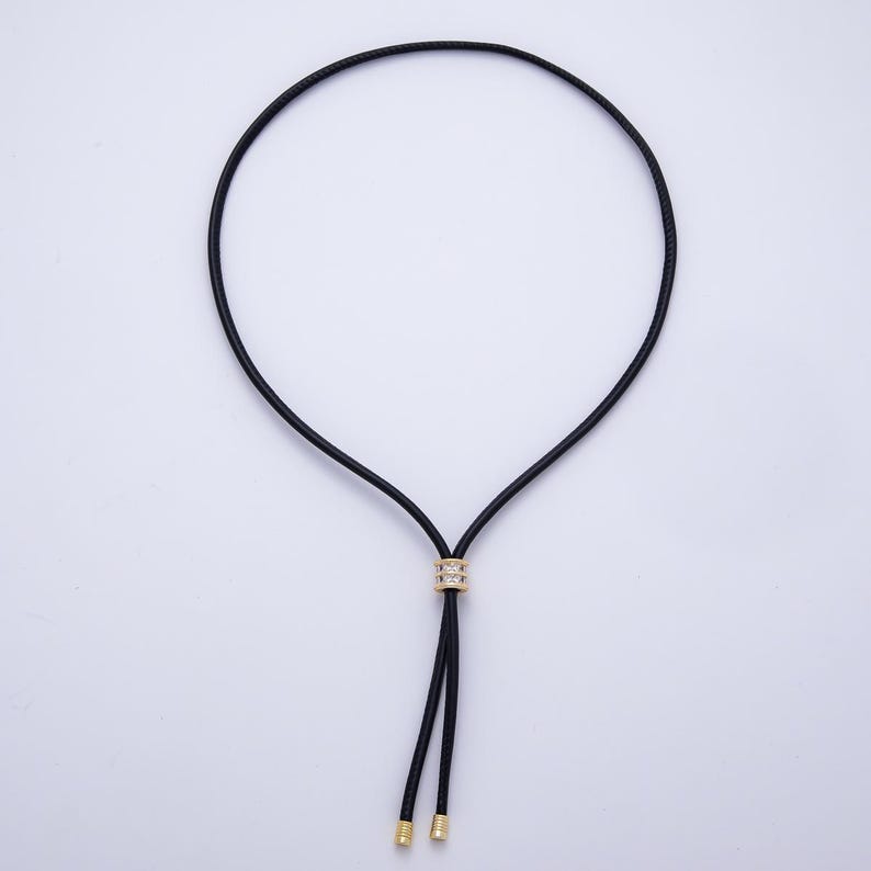 May include: A black cord necklace with a gold-colored central bead encrusted with small, clear stones. The necklace has two dangling black cords with gold-colored tips. The necklace is displayed against a white background.