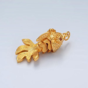 May include: A gold-coloured pendant shaped like a fish with a red gemstone eye. The pendant has intricate details and a textured surface.