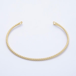 May include: A gold-colored, open cuff bracelet with a textured, geometric pattern. The bracelet has a slight curve and is open at the ends, with small loops. The jewelry is likely made of metal and is designed to be worn on the wrist.