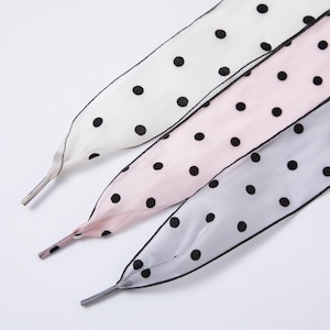 May include: Three shoelaces in white, pink, and gray, each adorned with black polka dots. The laces have a delicate, sheer appearance and are edged with black stitching. The tips are capped with a silver-colored metal.