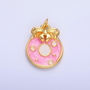 May include: A gold-colored pendant with a pink enamel donut design. The donut has a white center and is adorned with white dots. A gold bow sits atop the donut, and the pendant has a gold bail for attaching to a chain.