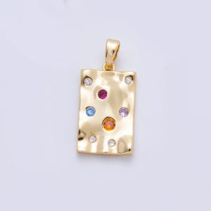 Pode incluir: Pingente retangular dourado com textura martelada. O pingente é adornado com uma variedade de pedras preciosas coloridas, incluindo um rubi central, pedras azuis, laranja e roxas, e pedras claras menores. Possui uma argola para corrente.