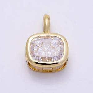 May include: A gold-toned pendant featuring a square-shaped, clear, faceted gemstone with rounded corners. The stone is set within a polished gold-colored metal frame that includes a vertical bail at the top for attachment to a chain or cord.