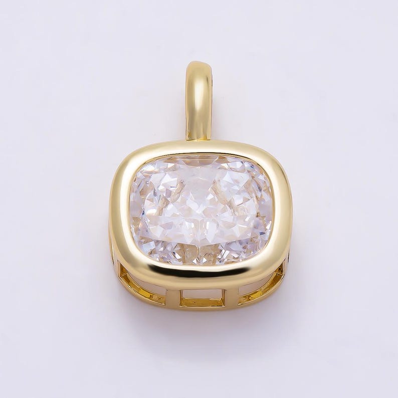 May include: A gold-toned pendant featuring a square-shaped, clear, faceted gemstone with rounded corners. The stone is set within a polished gold-colored metal frame that includes a vertical bail at the top for attachment to a chain or cord.