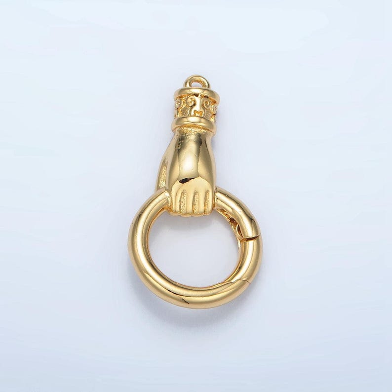 K&ouml;nnte beinhalten: Ein goldfarbener Verschluss mit einer Hand, die einen Ring h&auml;lt. Der Verschluss hat ein detailliertes Design auf der Oberseite und eine federbelastete &Ouml;ffnung am Ring. Das Gesamtdesign ist verziert und dekorativ.