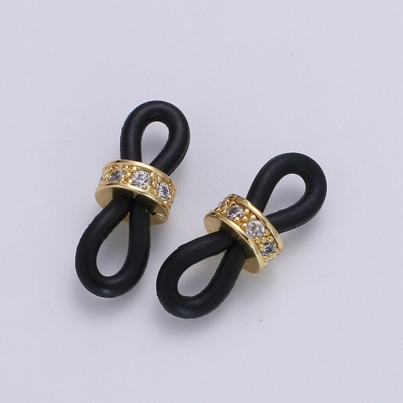 Puede incluir: Dos abalorios de bucle infinito de goma negra con detalles met&aacute;licos dorados y diamantes de imitaci&oacute;n transparentes.