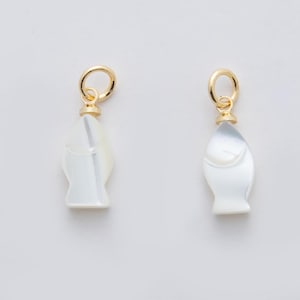 May include: Two fish-shaped charms. Each charm is white with a pearlescent sheen and a gold-colored loop at the top. The fish have a simple, stylized design, with a rounded body and a small tail. The charms are likely for jewelry.