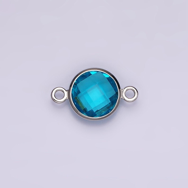 Puede incluir: Un conector plateado con una piedra preciosa redonda de color azul brillante. El conector tiene dos bucles a cada lado para sujetarlo a joyas.