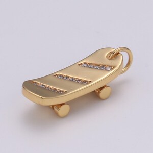 May include: A gold-toned skateboard charm with clear crystal accents. The charm has a loop at the top for attaching to a necklace or bracelet.
