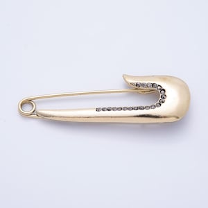 May include: A gold-colored safety pin with a row of small, dark-colored stones along the curved edge and the bottom edge. The pin is set against a white background.