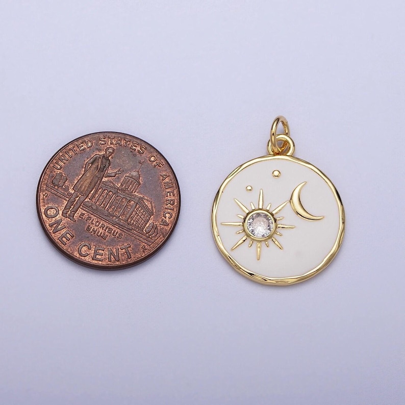 May include: Gold pendant charm with a white enamel background featuring a sun with a clear rhinestone center and a crescent moon. The pendant is approximately the size of a US penny.