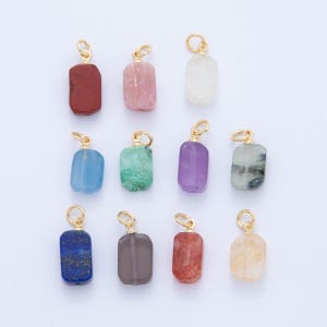 Może przedstawiać: Kolekcja wisiorków z kamieni szlachetnych w różnych kolorach, w tym czerwonym, różowym, niebieskim, zielonym, fioletowym i żółtym. Każdy prostokątny kamień ma na górze złotą pętlę do przymocowania do łańcuszka lub bransoletki. Wisiorki są wyświetlane na jasnym tle.