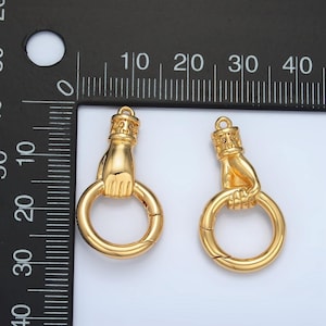 K&ouml;nnte beinhalten: Zwei goldfarbene Metallverschl&uuml;sse, jeweils mit einer Hand, die einen kreisf&ouml;rmigen Ring h&auml;lt. Die Verschl&uuml;sse haben ein detailliertes Design an der Hand und am oberen Teil. Der Hintergrund ist wei&szlig;.