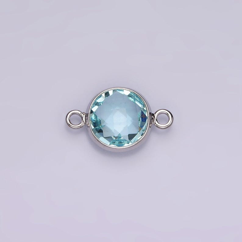 Puede incluir: Un conector plateado con una piedra redonda de color azul claro. El conector tiene dos bucles a cada lado para sujetarlo a joyas.