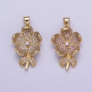 Puede incluir: Dos colgantes de flores de tono dorado con piedras de circonita cúbica transparentes y rosas. Los colgantes están detallados con un diseño de flor y un tallo.